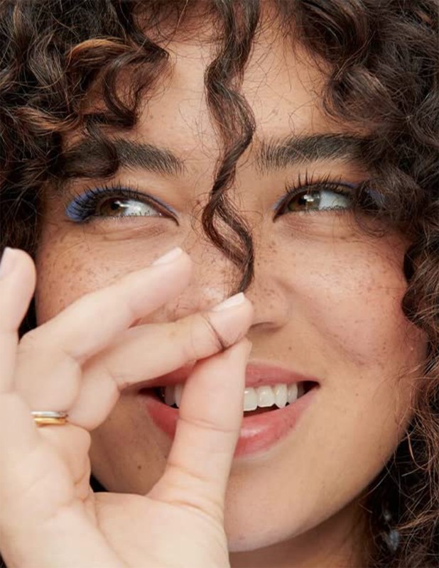 Portrait of a woman with curly brown hair and freckles smiling, holding a strand of hair between her fingers, with natural light.