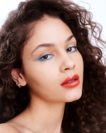 Close up portrait of a young woman with voluminous curly brown hair, blue winged eyeliner, and glossy red lipstick against a white background