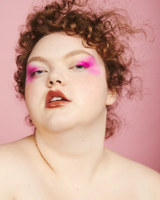 Close-up of a young adult female with fair skin and curly red hair, wearing bright pink eyeshadow and glossy lipstick, on a pink background.