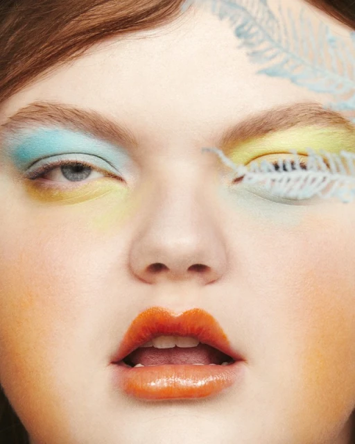 Close-up of a young woman's face with vibrant, creative makeup featuring colorful eyeshadow and glossy orange lips. Feather detail on face.