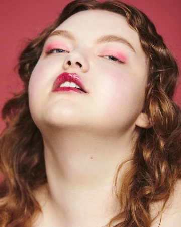 Close-up portrait of a woman with vibrant pink eyeshadow, glossy burgundy lipstick, and curly brown hair against a solid red backdrop.