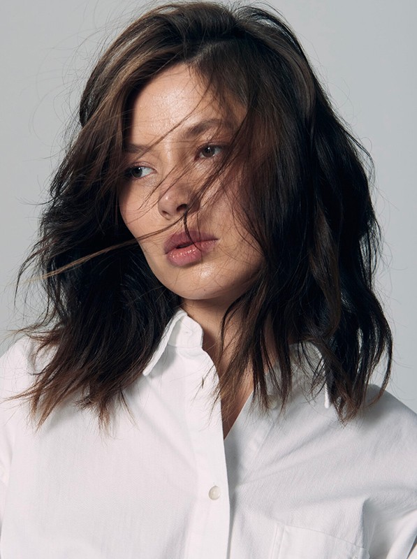 Close-up portrait of a woman with dark wavy hair and tousled strands across her face wearing a white collared shirt against a grey background.