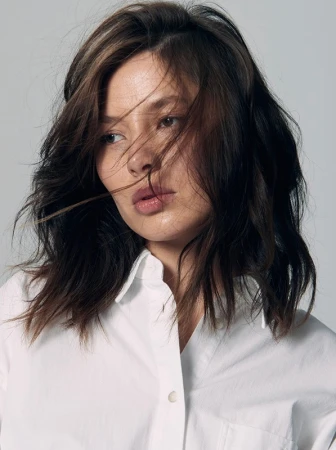 Close-up portrait of a woman with dark wavy hair and tousled strands across her face wearing a white collared shirt against a grey background.
