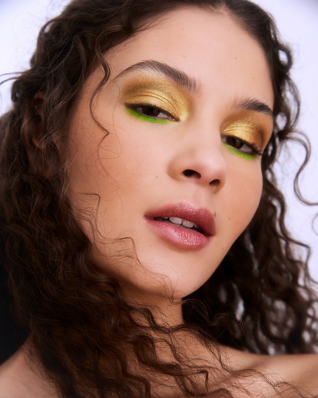 Close-up portrait of a young woman with golden eyeshadow and dark curly hair, posing in a photo studio with soft, diffused lighting.