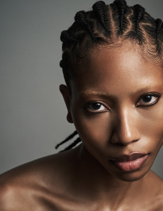 Close-up portrait of a Black woman with braided hair and dark eyeliner against a grey background.