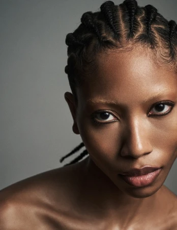 Close-up portrait of a Black woman with braided hair and dark eyeliner against a grey background.