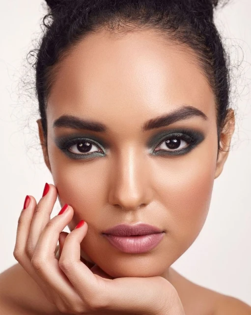 Close-up portrait of a woman with dark hair in a bun, green eyeshadow, red nail polish, and mauve lipstick against a white background.