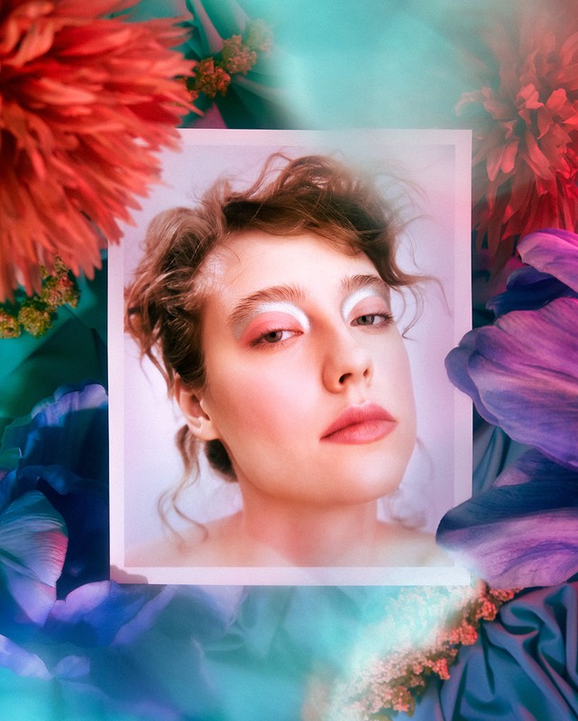 Close-up portrait of a woman with creative eye makeup, framed by colorful flowers in hues of red, purple, and teal with soft lighting.