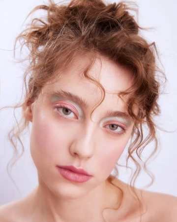 Close-up portrait of a woman with curly red hair, pink eyeshadow, and soft glossy makeup against a plain white background