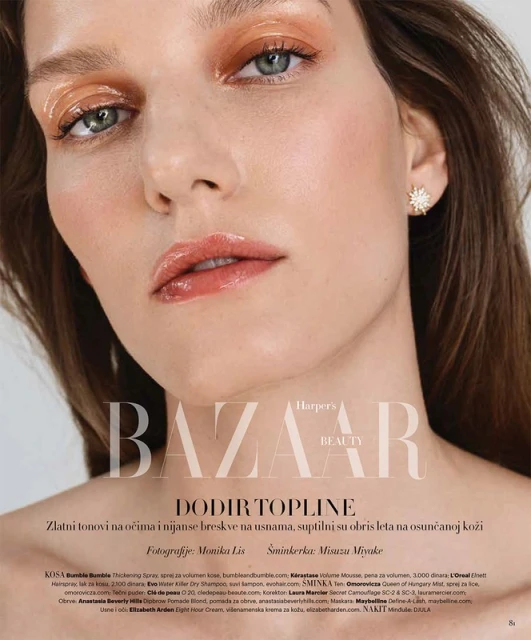 Close-up beauty shot of a young adult woman with glossy peach eye makeup and lips, fair skin, star-shaped earrings, and a Harper's Bazaar logo.