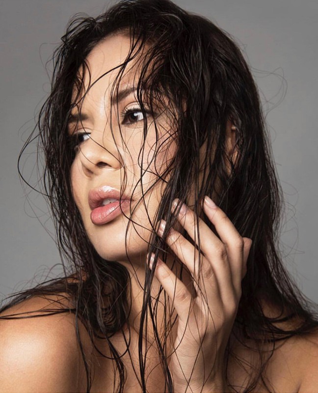 Close-up beauty portrait of a woman with wet dark hair touching her face against a neutral grey background