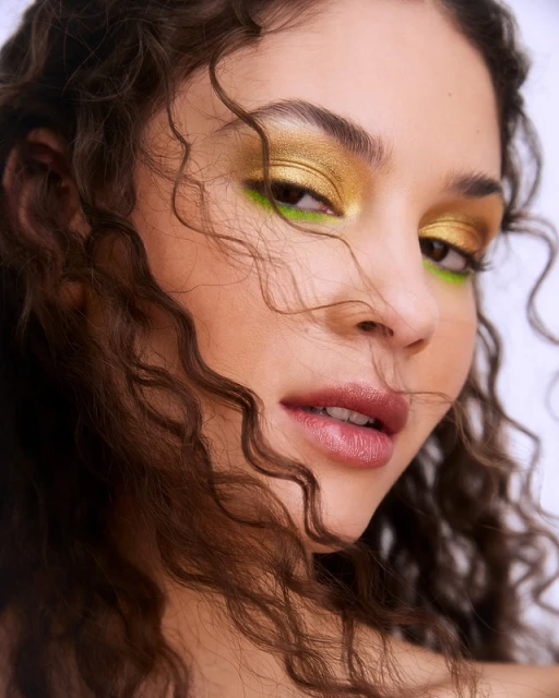 Close-up portrait of a young woman with curly brown hair and vibrant gold and green makeup against a bright white background.