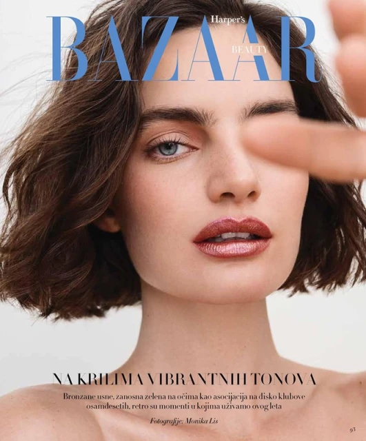 Close-up beauty shot of a brunette woman on a magazine cover with bronze lips and one eye winking, showcasing makeup and short wavy hair.
