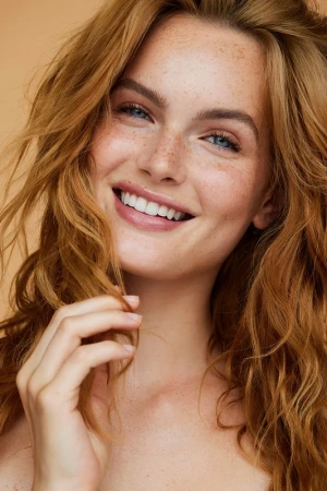 Close-up portrait of a smiling young woman with long wavy red hair and visible freckles against a neutral background
