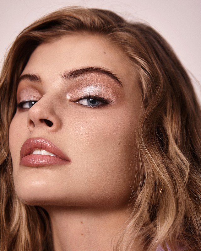 Close-up portrait of a fair-skinned adult woman with light brown hair, wearing shimmering eye makeup and glossy lips, against a soft pink backdrop.