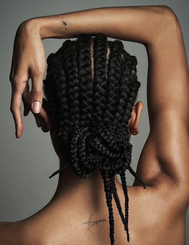 Back view of a Black woman with braided hair styled in a bun, arms raised, showcasing bare skin and a delicate shoulder tattoo.