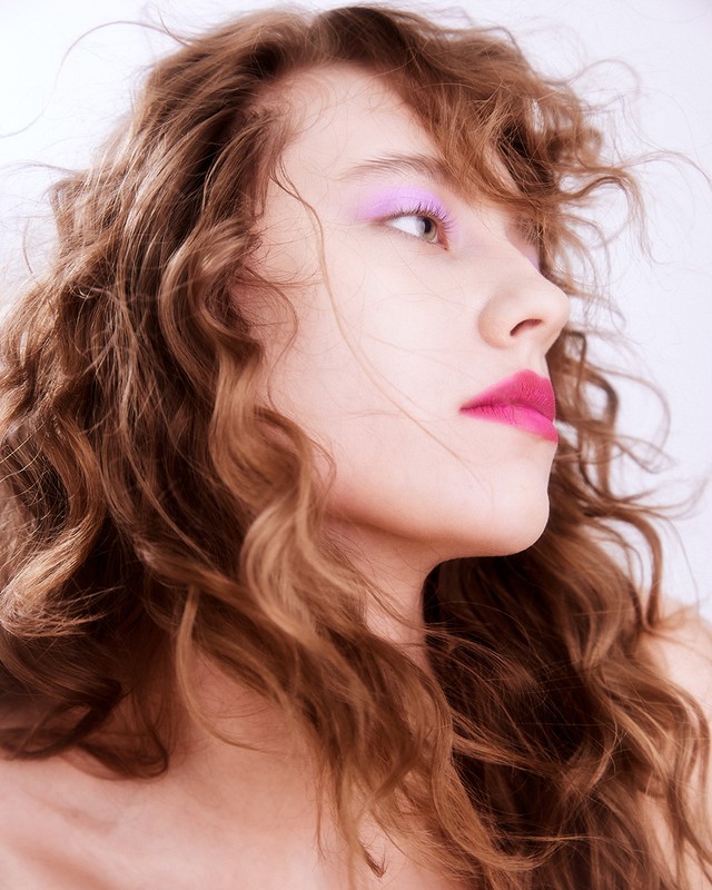 Profile portrait of a woman with curly brown hair and bright pink lipstick and lavender eyeshadow against a light background.