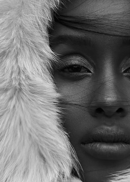 Close-up portrait of a Black woman with neutral expression, wearing makeup and a fur-trimmed hood in a high-contrast black and white studio shot.