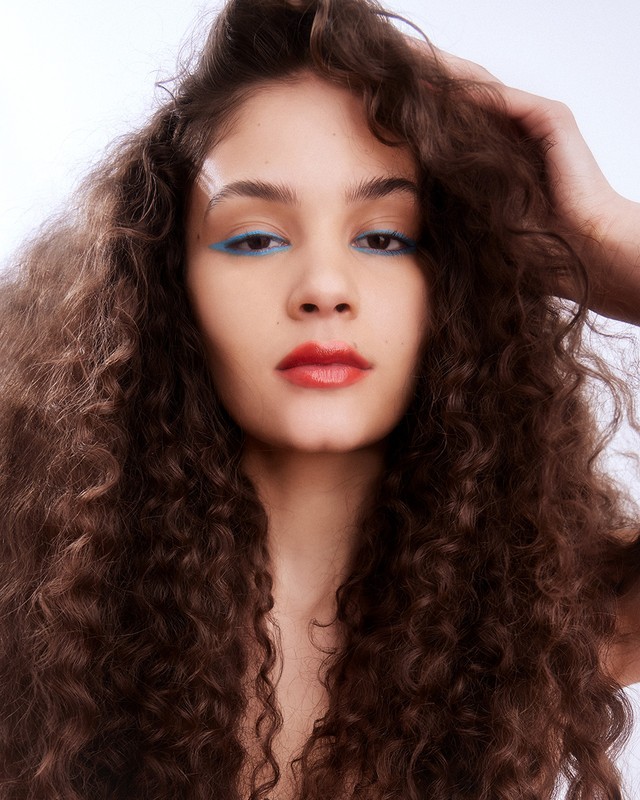 Close-up portrait of a woman with voluminous curly brown hair and blue eyeshadow posing against a white background