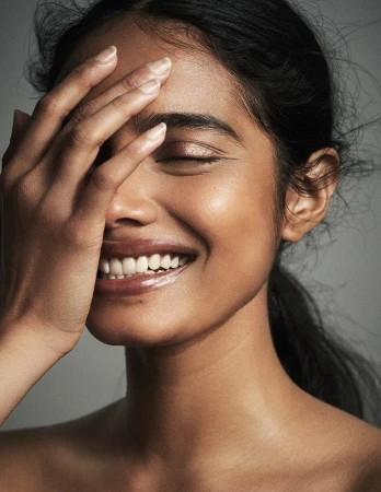 Close-up portrait of a smiling South Asian woman with her hand covering part of her face, styled by Destiny Venice.