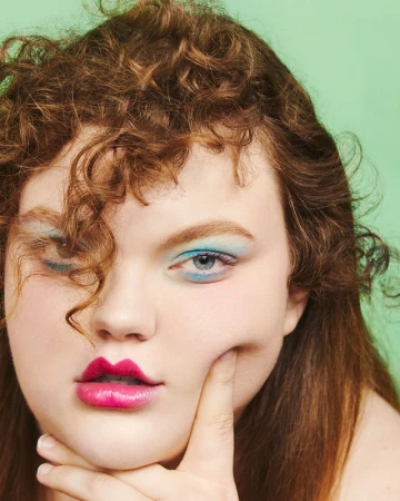 Close-up portrait of a woman with curly brown hair, blue eyeshadow, and bright pink lips against a solid green background.