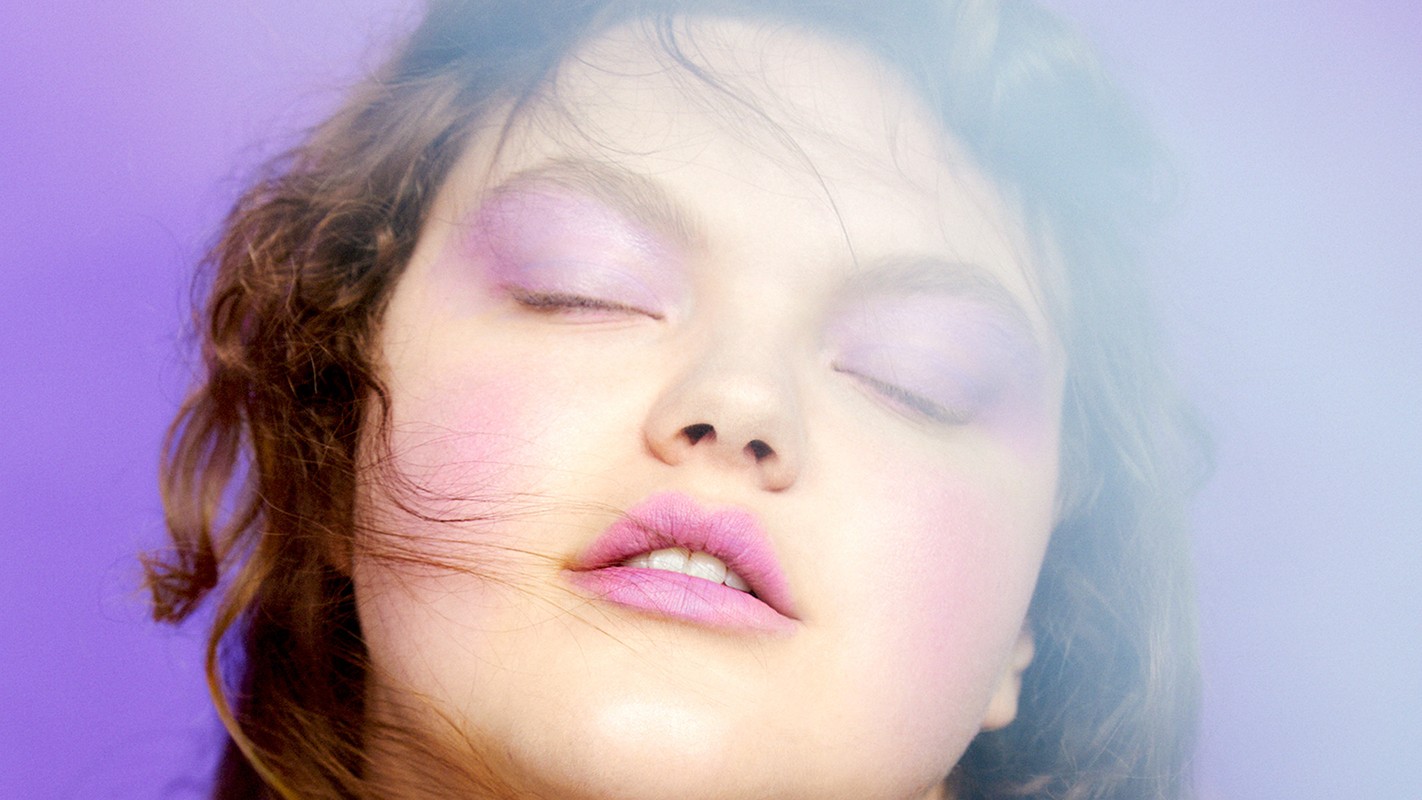 Close-up portrait of a woman with closed eyes, wearing lavender eyeshadow and pink lipstick, against a solid purple backdrop.