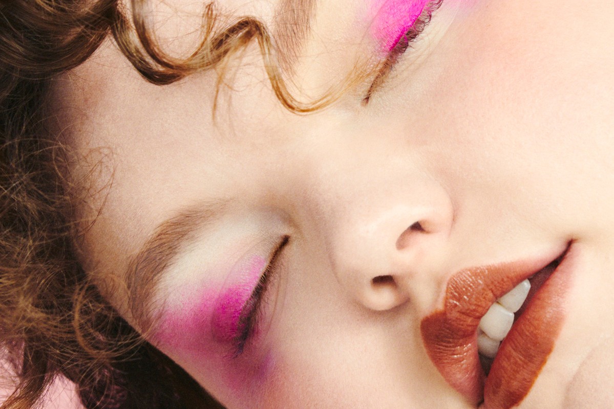 Close-up of a woman's face with pink eye makeup, brown curly hair, and glossy brown lips, showcasing a beauty and makeup aesthetic.
