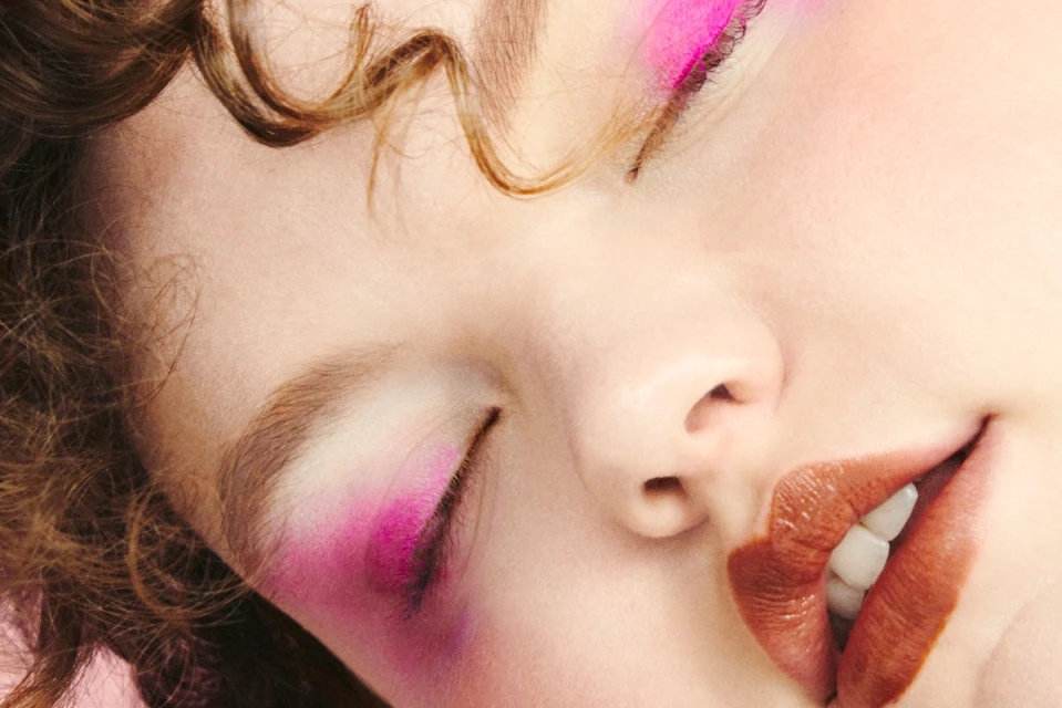 Close-up of a woman's face with pink eye makeup, brown curly hair, and glossy brown lips, showcasing a beauty and makeup aesthetic.
