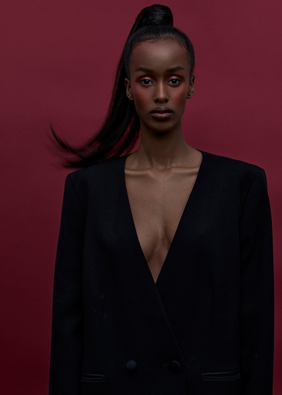 Portrait of a Black female model with a ponytail and open blazer against a deep red background in a studio setting. Fashion editorial.