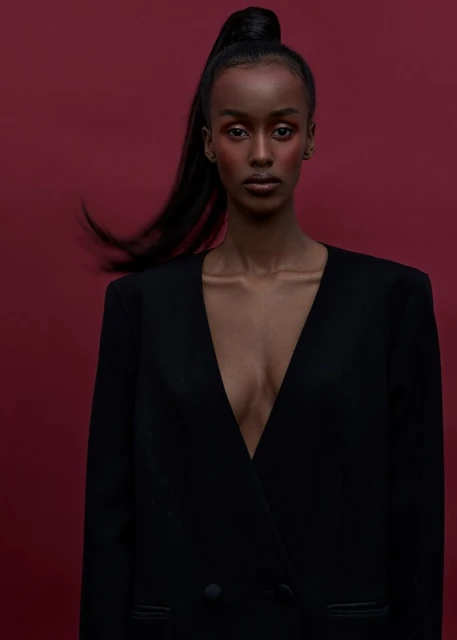 Portrait of a Black female model with a ponytail and open blazer against a deep red background in a studio setting. Fashion editorial.