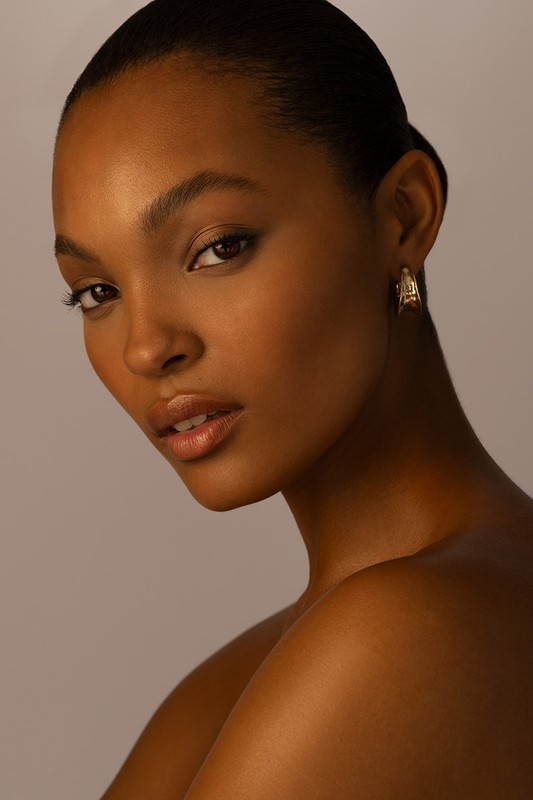Close-up portrait of a Black female model with slicked-back hair, wearing gold earrings against a soft, neutral background in a studio.