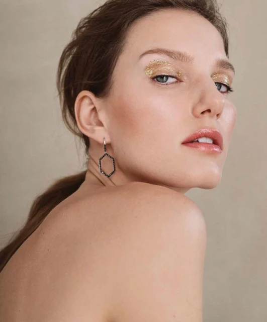Close-up portrait of a woman with gold glitter eyeshadow and glossy lips, wearing geometric earrings, against a neutral backdrop.