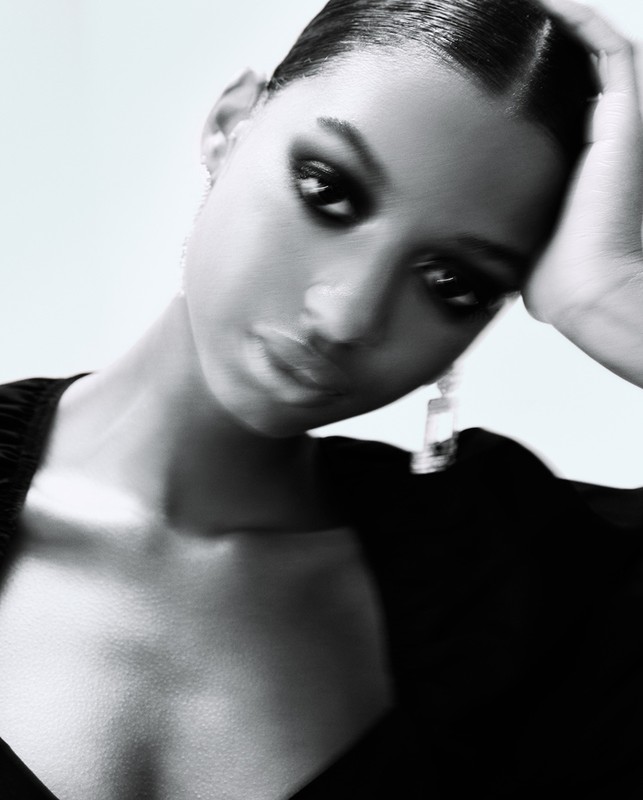 Close-up portrait of a Black female fashion model with dark smokey eyes and glossy lips, wearing dangling earrings, posing against a white background.