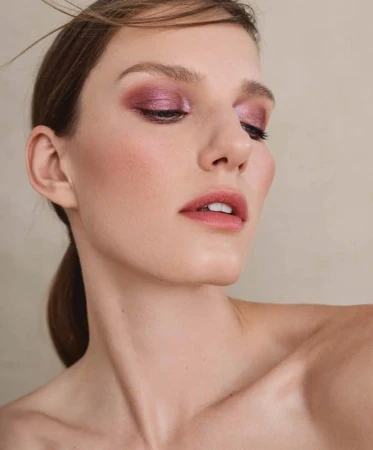 Close-up portrait of a woman with metallic pink eyeshadow and mauve lips against a neutral background.