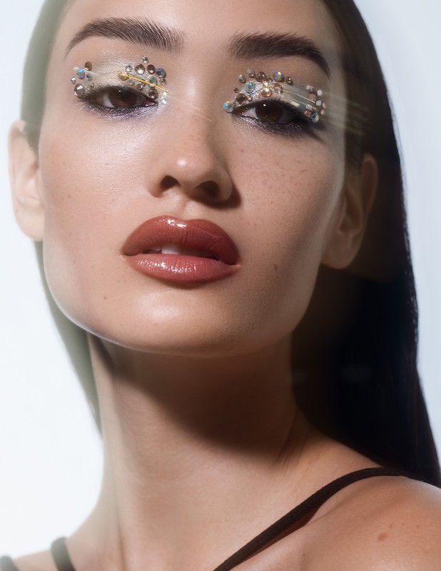 Close-up portrait of a young woman with jeweled eye makeup, glossy lips, and dark hair against a bright white background.