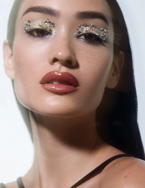 Close-up portrait of a young woman with jeweled eye makeup, glossy lips, and dark hair against a bright white background.