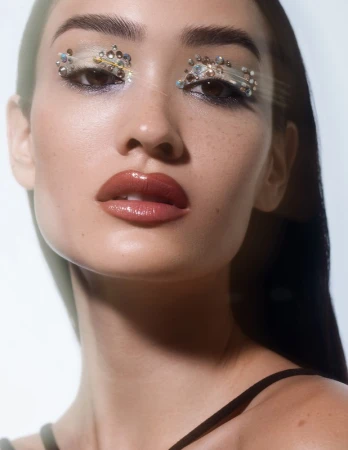 Close-up portrait of a young woman with jeweled eye makeup, glossy lips, and dark hair against a bright white background.