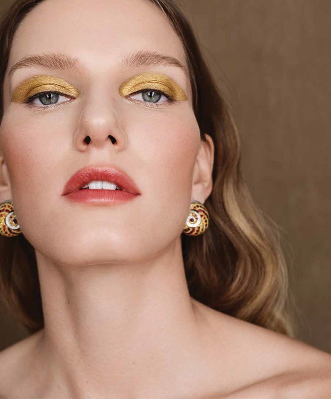 Close-up portrait of a blonde woman with gold eyeshadow and statement earrings against a neutral background, ideal for beauty or fashion content.