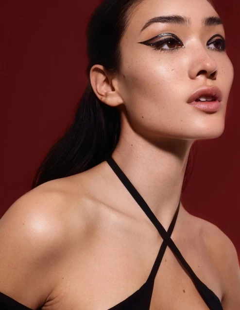 Close-up portrait of a woman with dramatic black eyeliner, dark hair pulled back, wearing a black halter top against a solid red background.