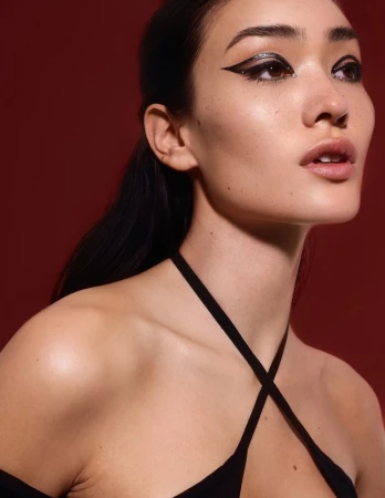 Close-up portrait of a woman with dramatic black eyeliner, dark hair pulled back, wearing a black halter top against a solid red background.