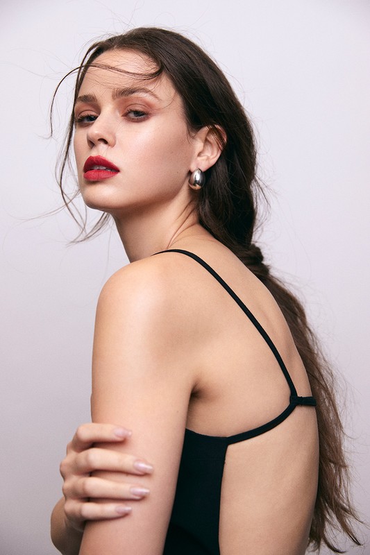 Portrait of a young woman with long brunette hair, wearing a black spaghetti strap top and silver hoop earrings, posing in a photo studio.