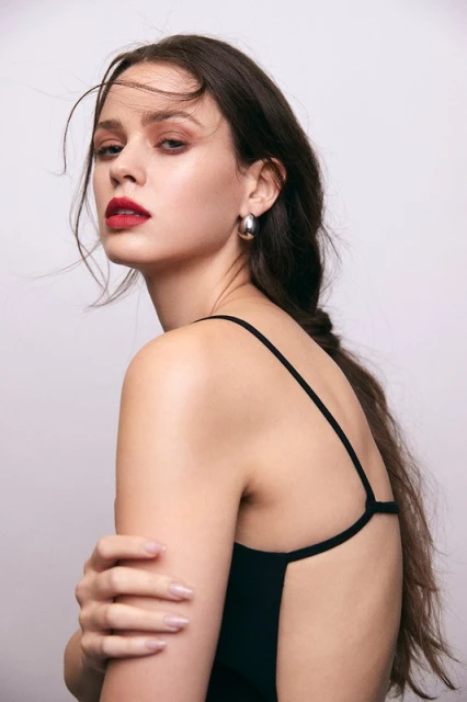 Portrait of a young woman with long brunette hair, wearing a black spaghetti strap top and silver hoop earrings, posing in a photo studio.