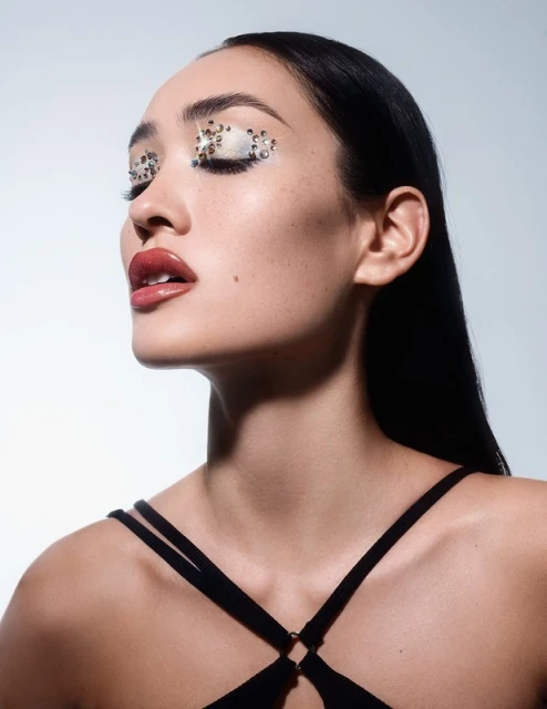 Close-up portrait of an adult woman with dark hair and a black top, wearing embellished eye makeup, set against a light blue background.