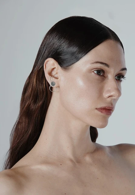 Close-up studio shot of an adult female model with smooth dark hair, minimal makeup, and round silver earrings against a plain background.