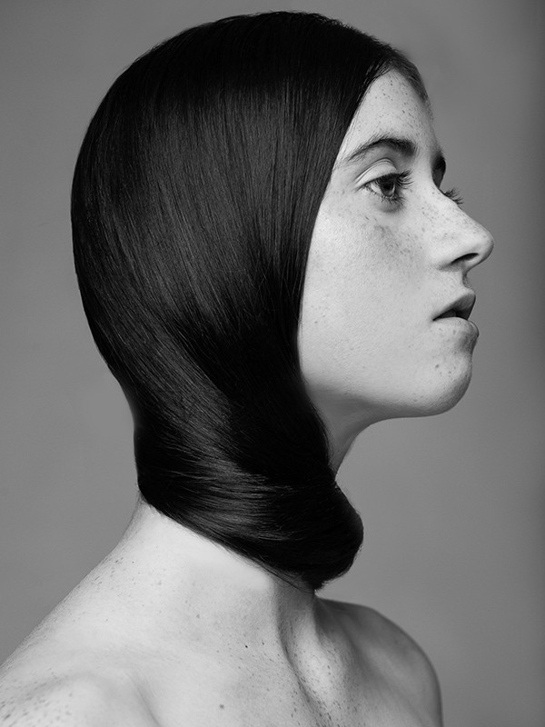 Striking black and white portrait of a bare-shouldered woman with slicked-back, sculptural black hair on a neutral grey background.