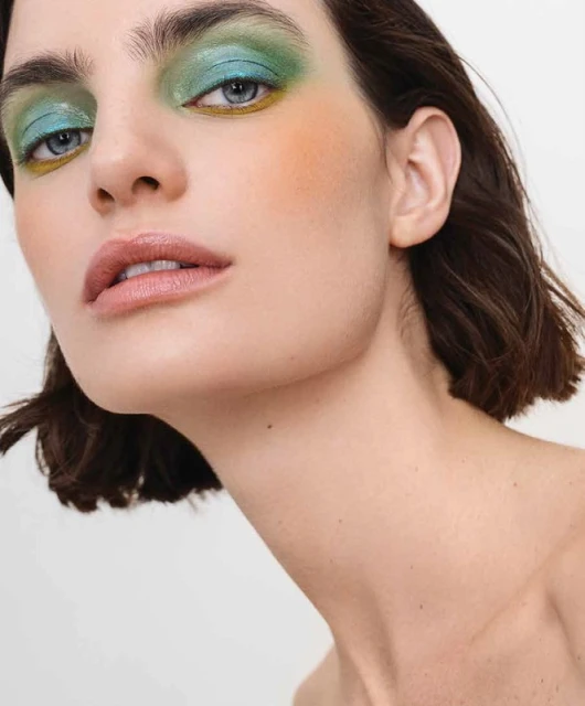 Close-up portrait of a young adult woman with blue eyes, light skin, and blue and green eyeshadow against a white background.