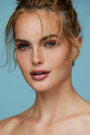 Close-up beauty portrait of a fair-skinned woman with heavy freckles, blue eyes, and loose blonde strands of hair against a blue background.