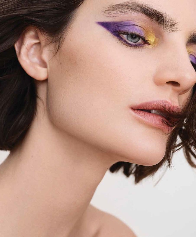 Close up beauty portrait of a woman with dramatic purple and yellow graphic eye makeup and dark short hair.