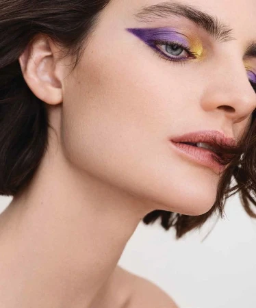 Close up beauty portrait of a woman with dramatic purple and yellow graphic eye makeup and dark short hair.