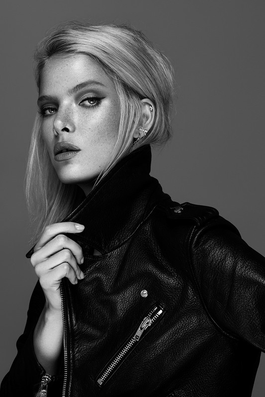 Black and white portrait of a blonde adult woman in a black leather jacket, posing in front of a grey background, looking at the camera.